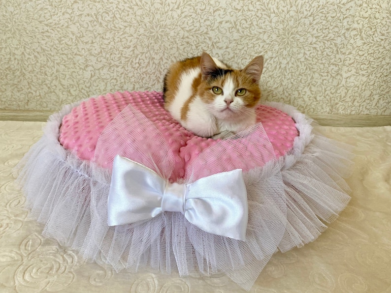 May include: A pink and white cat bed with a white tulle skirt and a white satin bow. A calico cat is sitting on the bed.