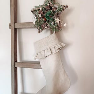 Pode incluir: Uma meia de Natal de linho bege com uma borda franjada pendurada em uma escada de madeira. Um enfeite de estrela de galho com ramos de pinheiro, pinhas e contas brancas está acima da meia.