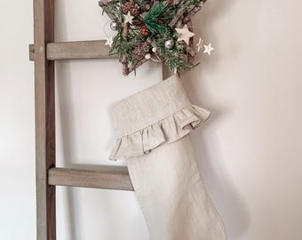 May include: A beige linen Christmas stocking with a ruffled edge hangs on a wooden ladder. A twig star ornament with pine sprigs, pinecones, and white beads is above the stocking.