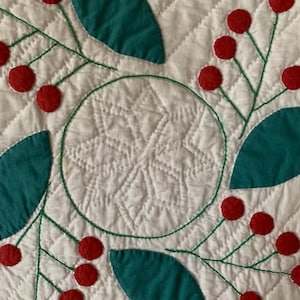May include: A white quilted fabric with a green and red design. The design features a circle with a starburst pattern in the centre, surrounded by green leaves and red berries.