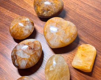 May include: Seven tumbled stones of citrine, a variety of quartz, in various shapes and sizes. The stones are a light yellow color with a translucent appearance. The stones are arranged on a wooden surface.