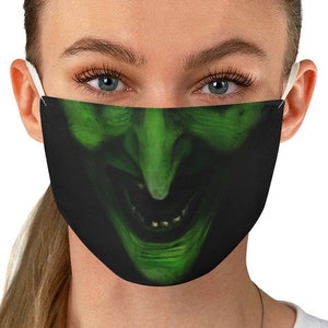 May include: A green and black face mask with a witch's face printed on it. The witch has a large nose and a wide smile.