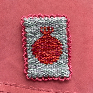 May include: A woven textile with a red pomegranate design on a light blue background, framed by a pink scalloped edge. The textile is rectangular and set against a pink fabric background.