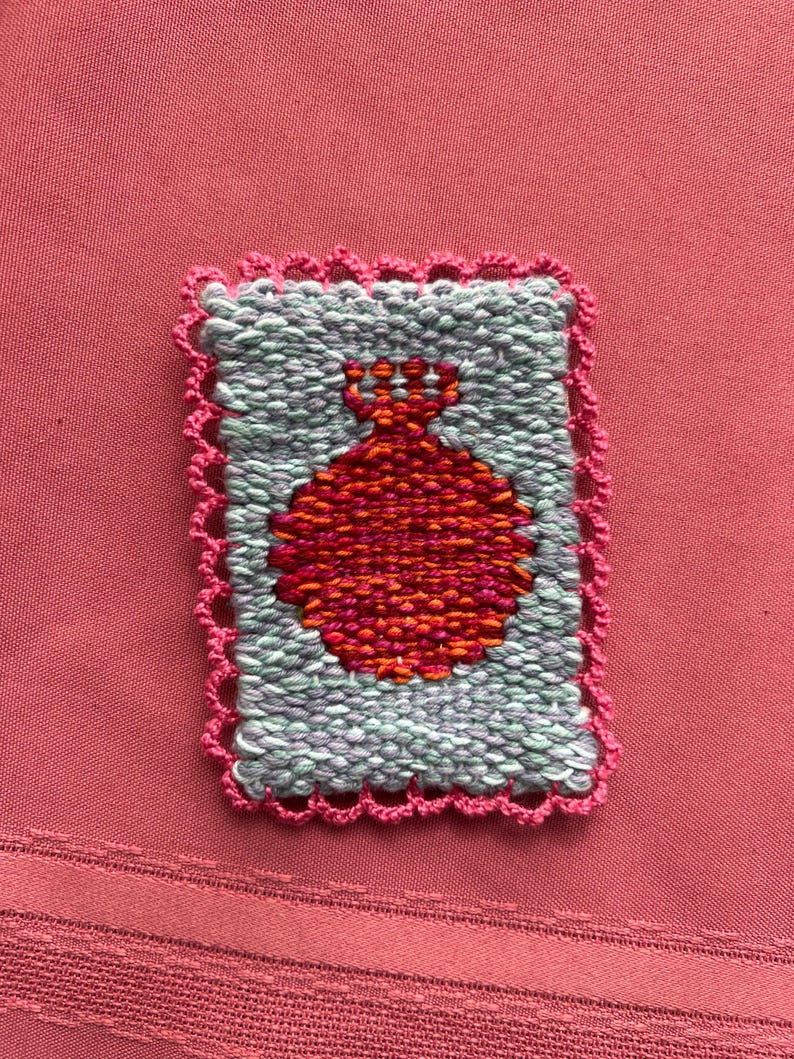May include: A woven textile with a red pomegranate design on a light blue background, framed by a pink scalloped edge. The textile is rectangular and set against a pink fabric background.
