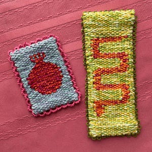 May include: Two hand-woven textile pieces. One is rectangular with a red design on a blue background, framed with pink. The other is a long, narrow rectangle with an orange design on a green background. Both are on a pink surface.