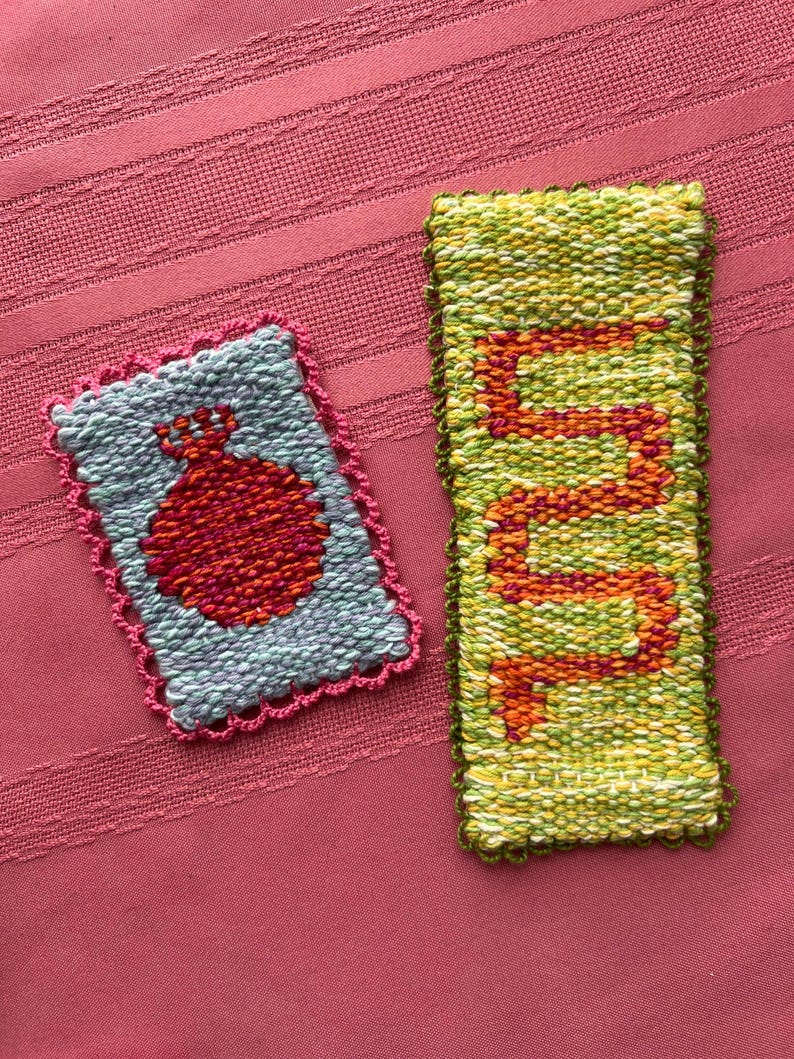 May include: Two hand-woven textile pieces. One is rectangular with a red design on a blue background, framed with pink. The other is a long, narrow rectangle with an orange design on a green background. Both are on a pink surface.