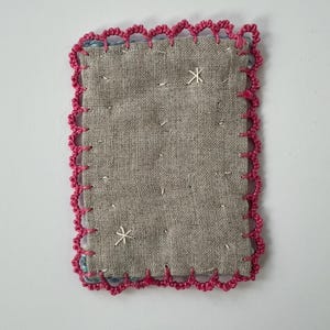 May include: A small, rectangular textile patch with a gray linen-like fabric center. The edges are trimmed with a bright pink, scalloped crochet border. Two white embroidered starburst designs are visible on the surface.