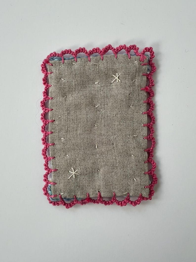 May include: A small, rectangular textile patch with a gray linen-like fabric center. The edges are trimmed with a bright pink, scalloped crochet border. Two white embroidered starburst designs are visible on the surface.
