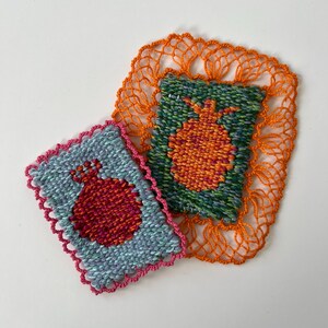 May include: Two handmade textile coasters. One is light blue with a red pomegranate design and a pink scalloped edge. The other is green with an orange pomegranate design and an orange lace border. The coasters are on a white background.