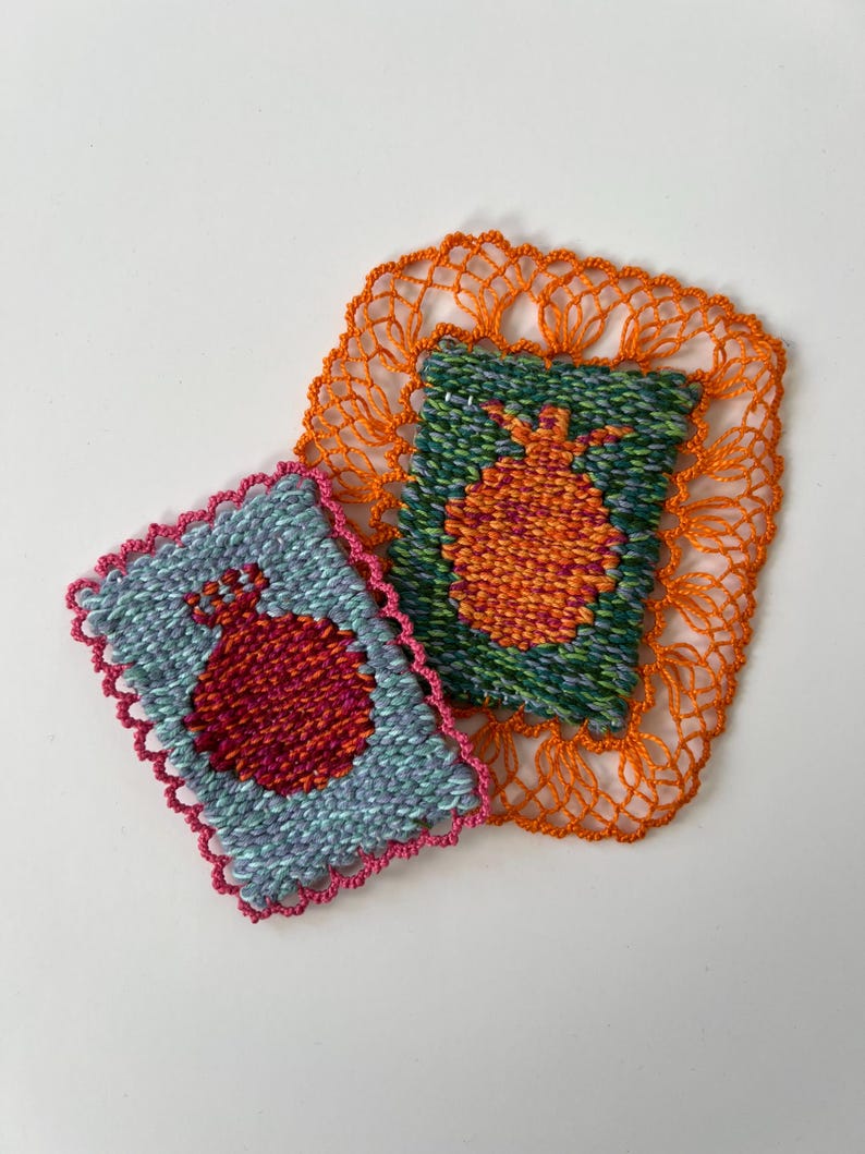 May include: Two handmade textile coasters. One is light blue with a red pomegranate design and a pink scalloped edge. The other is green with an orange pomegranate design and an orange lace border. The coasters are on a white background.
