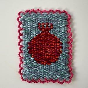 May include: Handwoven textile art piece featuring a red pomegranate design on a teal and gray background. The artwork is framed with a scalloped pink border, showcasing a textured, handcrafted aesthetic. This unique piece is suitable for home decor.