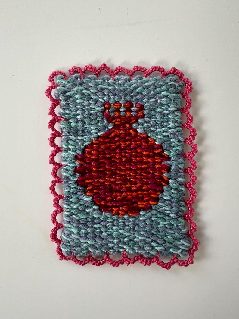 May include: Handwoven textile art piece featuring a red pomegranate design on a teal and gray background. The artwork is framed with a scalloped pink border, showcasing a textured, handcrafted aesthetic. This unique piece is suitable for home decor.