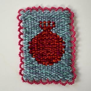 May include: Handwoven textile art piece featuring a red pomegranate design on a teal and gray background. The artwork is framed with a pink, scalloped border, showcasing a unique texture and color palette.