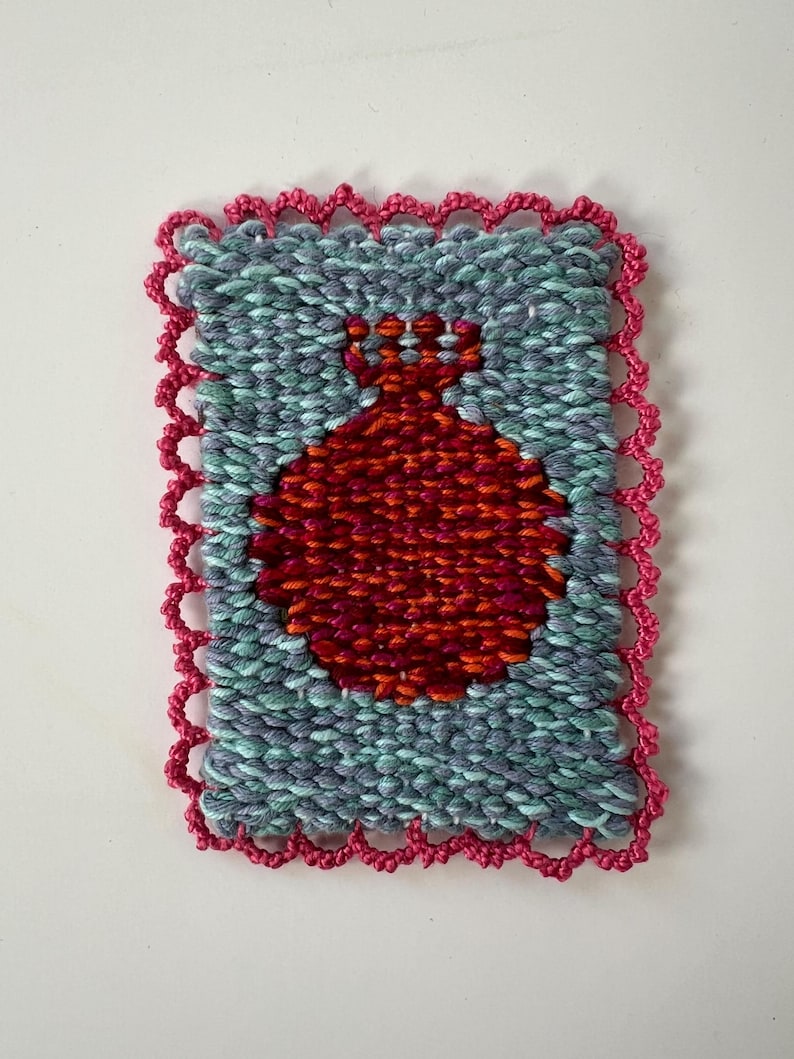 May include: Handwoven textile art piece featuring a red pomegranate design on a teal and gray background. The artwork is framed with a pink, scalloped border, showcasing a unique texture and color palette.