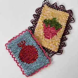 May include: Two handmade woven coasters. One features a red pomegranate on a blue background with a pink scalloped edge. The other has a pink strawberry with green leaves on a yellow background, with a dark brown scalloped edge.