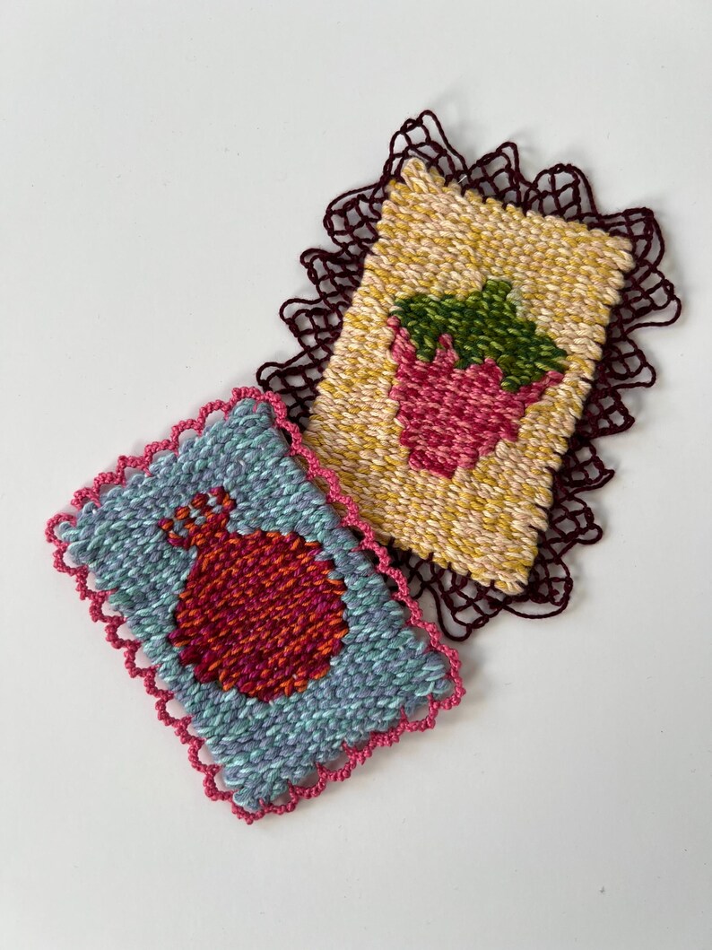 May include: Two handmade woven coasters. One features a red pomegranate on a blue background with a pink scalloped edge. The other has a pink strawberry with green leaves on a yellow background, with a dark brown scalloped edge.