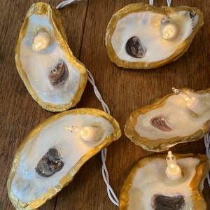 Puede incluir: Una cadena de luces con conchas de ostras pintadas de oro, cada una con una pequeña bombilla en su interior. Las conchas están unidas por un cable blanco.