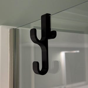 Puede incluir: Un gancho de plástico negro con dos puntas, diseñado para colgar objetos en una superficie de vidrio.