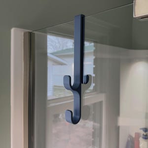 Könnte beinhalten: Ein dunkelgrauer Duschhaken für die Tür. Der Haken hat ein kaktusartiges Design mit drei gebogenen Haken. Er hängt an einer Glas-Duschwand.