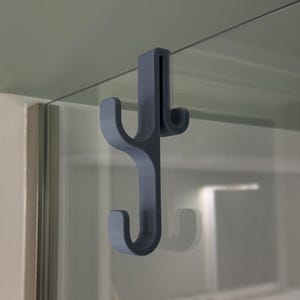 Puede incluir: Un gancho de plástico gris oscuro con dos ganchos en el extremo. El gancho está diseñado para colgarse sobre una puerta o estante de vidrio.