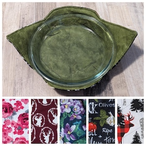May include: A clear glass pie dish is nestled within a green fabric pie cozy. The cozy features a green interior and a patterned edge. Below are fabric swatches with floral, deer, and food-themed designs.