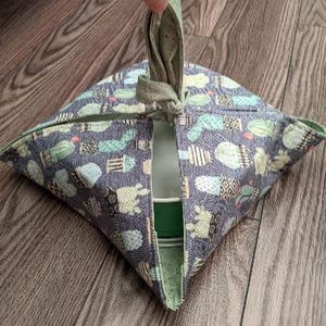 May include: A fabric bowl cozy featuring a repeating pattern of various green and white cacti on a dark gray background. The square-shaped quilted holder has four folded corners that meet at a central knotted handle, partially revealing a green bowl.
