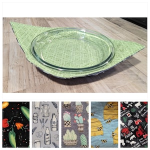 Peut inclure: Un plat à tarte en verre transparent repose sur un dessous de plat en tissu vert matelassé. Le dessous de plat a une forme de feuille avec un motif vert clair. Le fond est une surface en bois clair.