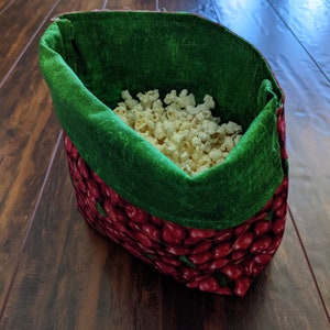 May include: A fabric bowl filled with white popcorn. The exterior features a repeating pattern of red cherries with green leaves on a dark background, while the interior is lined with a solid, textured green fabric that folds over the top edge.