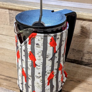Könnte beinhalten: Eine French-Press-Kaffeebereiter aus Glas mit schwarzem Kunststoffdeckel, Griff und Boden. Die Glaskaraffe ist mit einer Stoffhülle umgeben, die mit einem Muster aus leuchtend roten Kardinalvögeln auf grauen und weißen Birkenstämmen bedruckt ist.