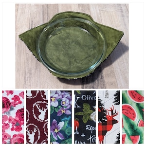 May include: A green fabric hot pad with a black and white trim, designed to hold a glass pie plate. The hot pad is made of a quilted fabric and has a soft, plush texture.