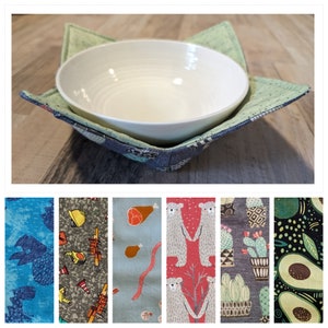 Könnte beinhalten: Eine weiße Keramikschüssel sitzt in einem grünen und blauen Stoffschüsselwärmer. Der Wärmer hat verschiedene Muster, darunter Baufahrzeuge, Tiere und geometrische Formen.