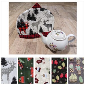 Puede incluir: Un calienta-tetera de cuadros rojos y negros con fondo blanco que representa una escena de bosque de invierno con ciervos y árboles. El calienta-tetera está sobre una superficie de madera junto a una tetera blanca con diseños florales.