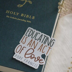 May include: A white sticker with the text "Educating is an act of love" in black and brown lettering. The text "Pope Francis" is written in smaller black lettering below the main text. The sticker is placed on a dark green book with the words "Holy Bible" in gold lettering.