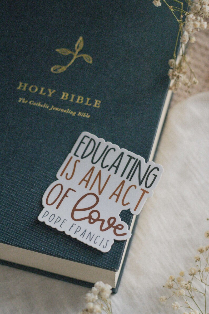 May include: A white sticker with the text "Educating is an act of love" in black and brown lettering. The text "Pope Francis" is written in smaller black lettering below the main text. The sticker is placed on a dark green book with the words "Holy Bible" in gold lettering.
