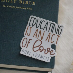 May include: A white sticker with the text "Educating is an act of love" in black and brown lettering. The text "Pope Francis" is written in black below the quote.