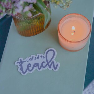 Pu&ograve; includere: Una candela accesa in un bicchiere color pesca &egrave; appoggiata su un quaderno verde chiaro. Un adesivo con le parole "called to teach" &egrave; sul quaderno. Un vaso con fiori &egrave; sullo sfondo.