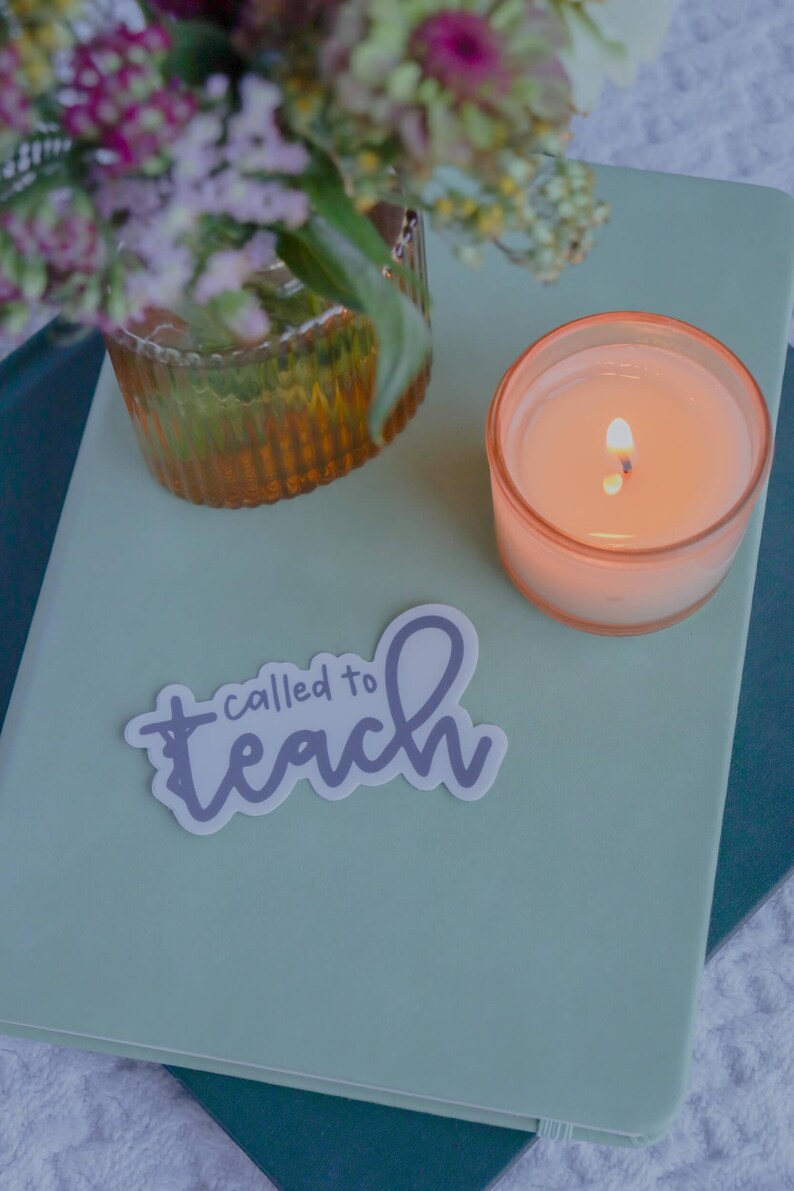 Pu&ograve; includere: Una candela accesa in un bicchiere color pesca &egrave; appoggiata su un quaderno verde chiaro. Un adesivo con le parole "called to teach" &egrave; sul quaderno. Un vaso con fiori &egrave; sullo sfondo.