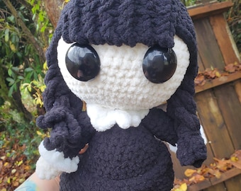 May include: A handmade crochet doll with a black dress, black hair in braids, and large black eyes. The doll has a white face and collar, and white trim on the dress and shoes. The doll is a soft, plush toy.