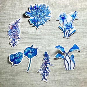 May include: A collection of blue floral stickers in various designs, including a chrysanthemum, daffodils, and other botanical illustrations. The stickers are hand-drawn with watercolor-style shading and are arranged on a light wood-grain surface.