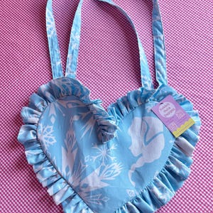 May include: A light blue heart-shaped tote bag with white snowflake and silhouette designs. The bag has ruffled edges and matching straps. A tag with the text "Rabbit Hole Designs" is attached.
