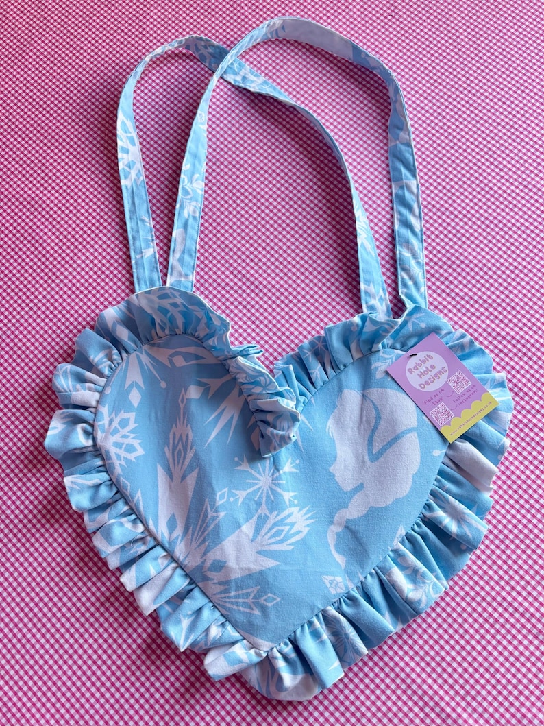 May include: A light blue heart-shaped tote bag with white snowflake and silhouette designs. The bag has ruffled edges and matching straps. A tag with the text "Rabbit Hole Designs" is attached.