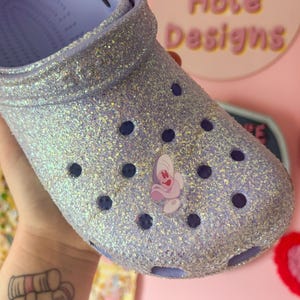May include: A lavender glitter-covered clog with a butterfly charm. The shoe has dark blue ventilation holes and a light purple interior. The words "Rabbit Hole Designs" are visible in the background.