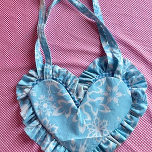 May include: A heart-shaped tote bag in light blue with white floral patterns. The bag features ruffled edges and matching straps. The background is a pink and white checkered pattern.