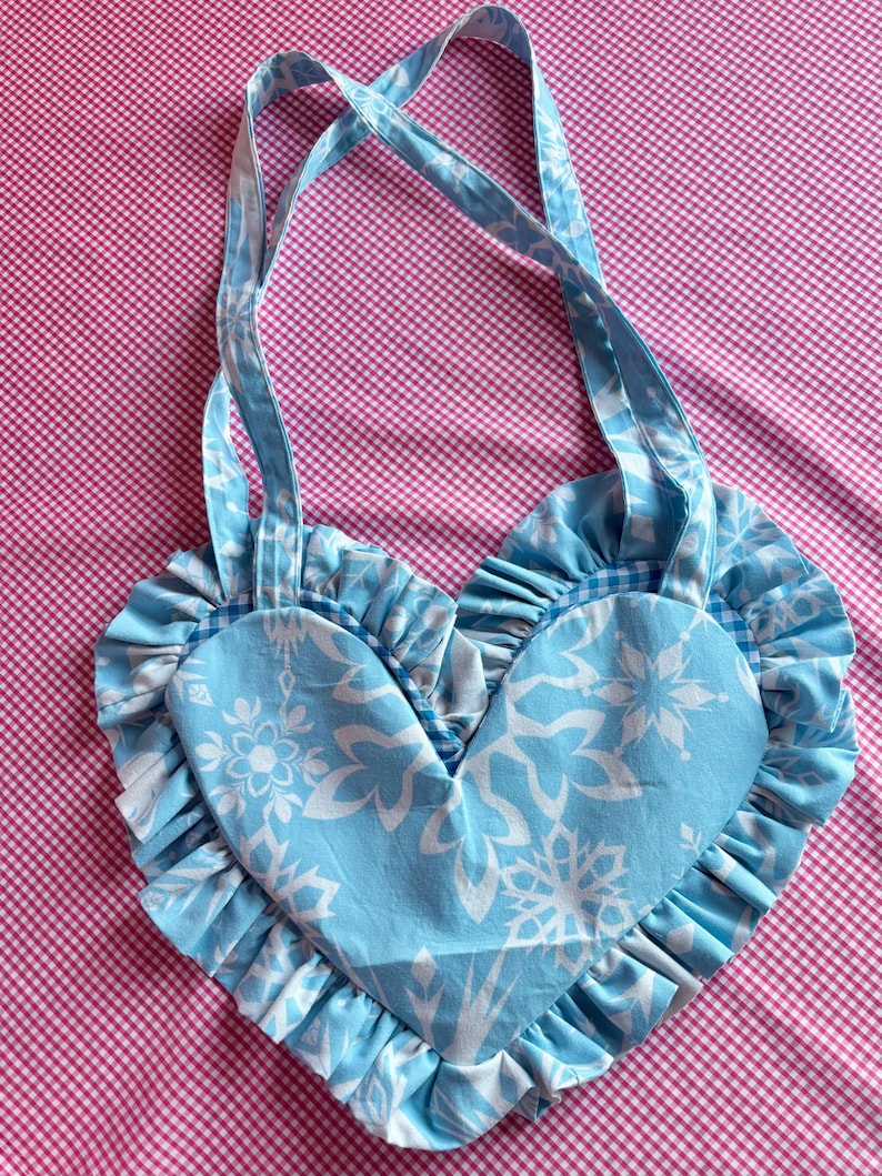 May include: A heart-shaped tote bag in light blue with white floral patterns. The bag features ruffled edges and matching straps. The background is a pink and white checkered pattern.