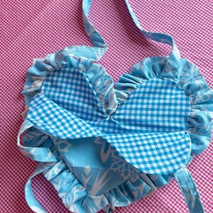May include: A heart-shaped, handmade bag with a blue and white floral pattern and a blue gingham pattern. The bag has a ruffled edge and long straps for carrying. The background is a pink and white checkered pattern.