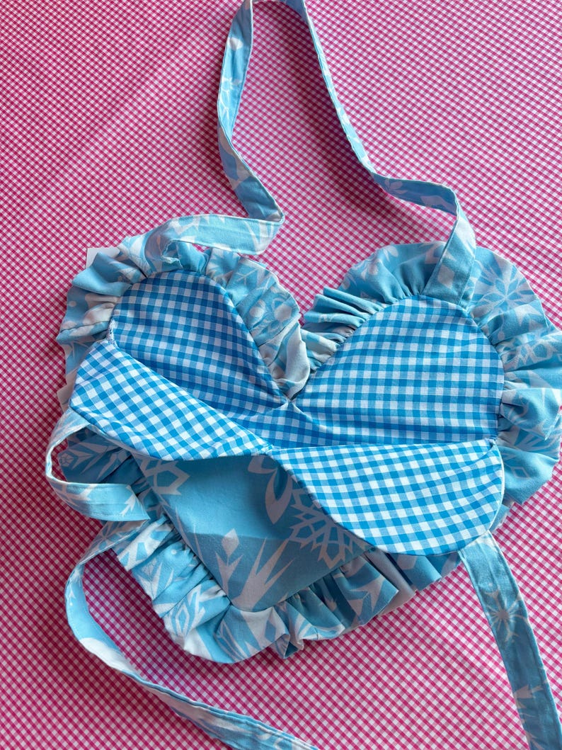 May include: A heart-shaped, handmade bag with a blue and white floral pattern and a blue gingham pattern. The bag has a ruffled edge and long straps for carrying. The background is a pink and white checkered pattern.