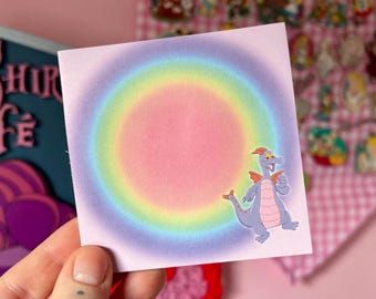 以下が含まれることがあります： 虹色のグラデーションの背景を持つ正方形の薄いピンク色のステッカー。漫画のドラゴンのキャラクターが右側にいます。ステッカーは手で持たれています。