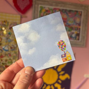 May include: A blue sticky note with a white cloud pattern and a small illustration of a house with balloons floating in the sky.
