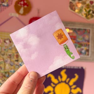May include: A purple card with a cartoon image of a green chameleon holding a lantern with a sun design. The lantern is floating in a sky with white clouds.
