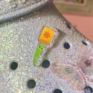 May include: A light blue shoe with a glittery finish. A clear charm with a green chameleon and a yellow sun with the text "You're Always With Me" is attached to the shoe. A clear butterfly charm is also attached to the shoe.